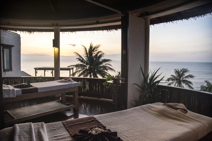 Open-air luxury spa treatment room with ocean view at sunset, featuring massage tables and tropical surroundings.