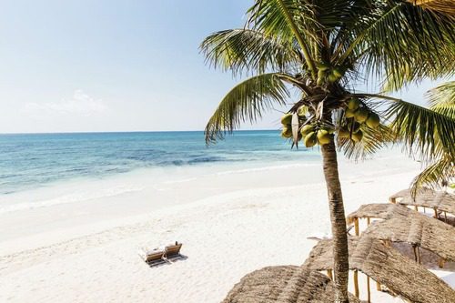 Tranquil luxury beach in Tulum, Mexico, with white sand, turquoise waters, palm trees, and shaded cabanas under a clear blue sky.