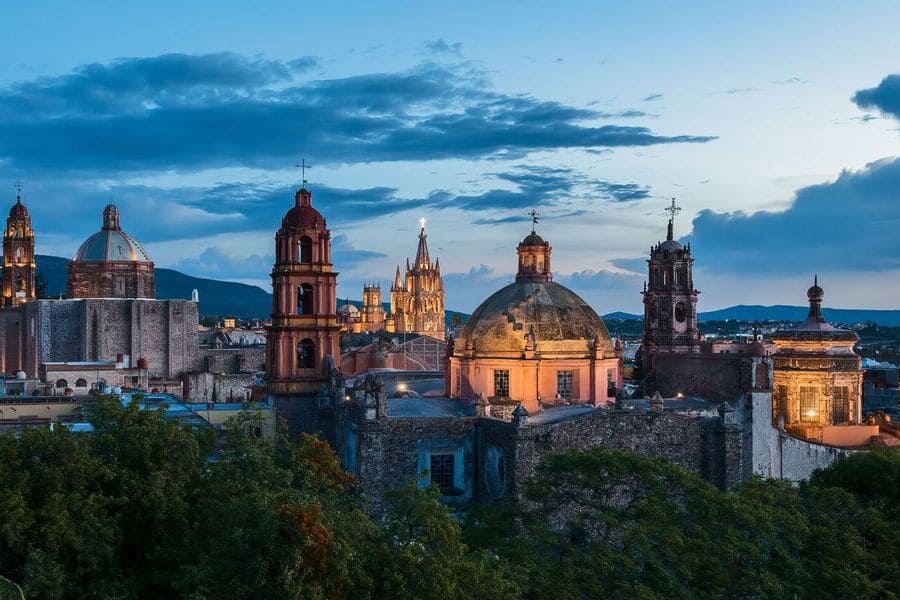 Historic skyline of San Miguel de Allende, Mexico at sunset, with illuminated colonial churches and dramatic evening skies.