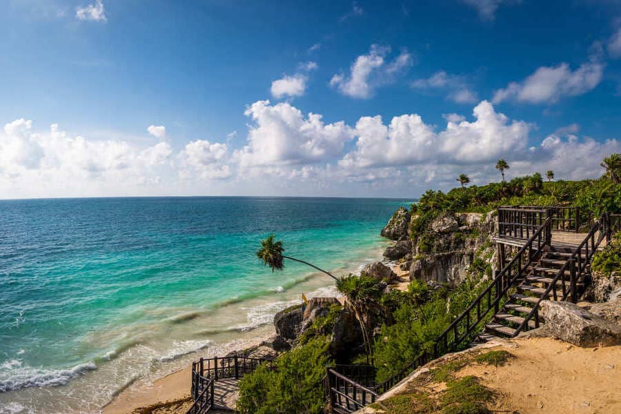 Scenic coastal view of Tulum’s white-sand beach and turquoise waters, with a wooden walkway leading to the shore.