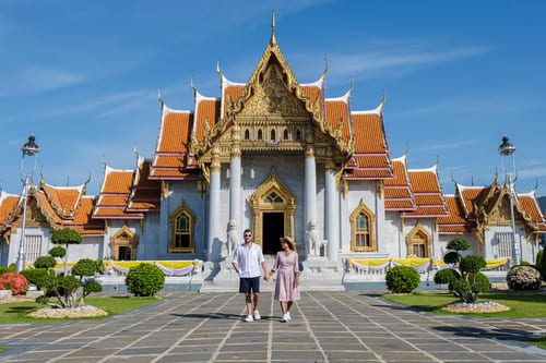 A couple walking in front of Wat Benchamabophit, the ornate marble temple in Bangkok, Thailand, under a bright blue sky.