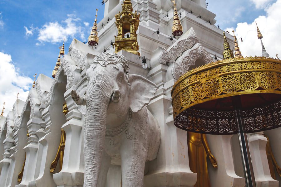 White elephant statue at the base of an ornate white and gold Buddhist temple in Thailand, under a bright blue sky with scattered clouds.